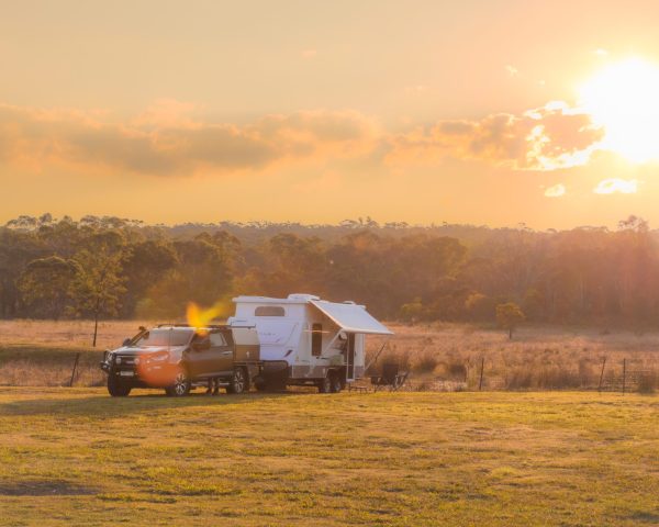 A camper trailer and car parked in an open field during sunset in Capertee Valley, with trees and a glowing sky in the background.